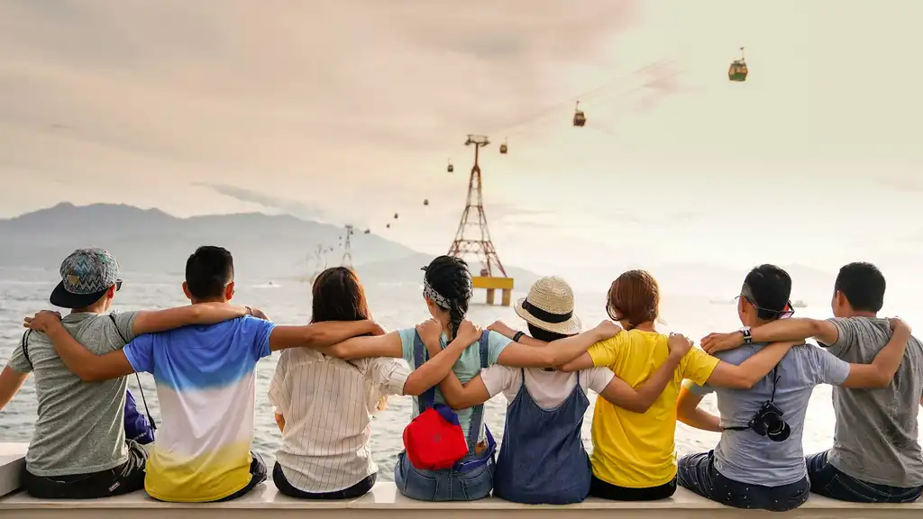 Group of people with arms over each other's shoulders sitting overlooking water looking at a mountain in the distance and funicular over water